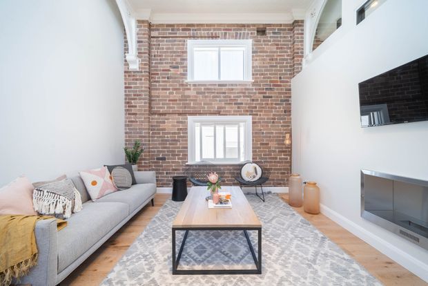 Bright double-height living room with a red exposed-brick feature wall, white decorative cornices and trim, timber flooring, a large patterned rug, and a pale grey sofa. A black-framed timber coffee table sits centrally, while a wall-mounted television and built-in fireplace occupy the white wall. High-level glazing and an upper opening emphasize the vertical volume.