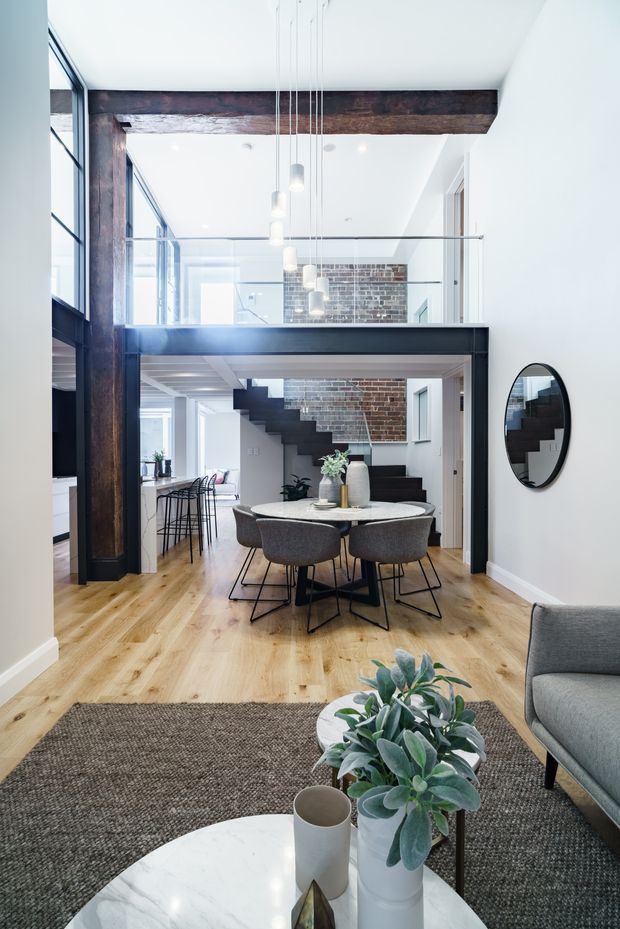 Bright open-plan interior with a dining area in the foreground, a living area partly visible at right, and a kitchen/bar zone to the left. The space features high ceilings, exposed dark timber beams, exposed brickwork, black steel-framed glazing and balustrades, a mezzanine level, and a contemporary floating staircase. Light timber flooring, neutral upholstered dining chairs, pendant lights, and indoor plants create an airy, refined industrial character.
