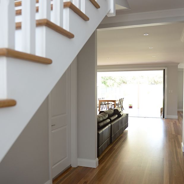 Bright transitional home interior viewed from a hallway, with a white staircase and warm timber handrails in the foreground, paneled doors, polished timber flooring, and an open-plan living area beyond. A dark leather sofa and dining table sit near large glazed doors opening to an outdoor area.