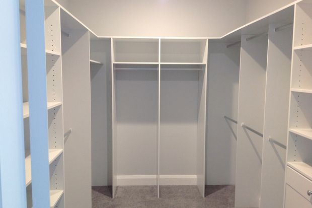A bright, built-in walk-in wardrobe with white shelving, hanging sections, drawers, and vertical storage units arranged along multiple walls. The room has grey carpet flooring and a simple white ceiling, with a clean, functional, minimalist fit-out.