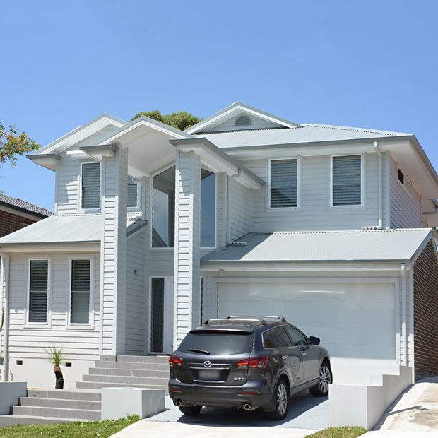 A bright exterior view of a substantial multi-storey suburban home with white horizontal weatherboard cladding, multiple gabled roof sections, louvred windows, a covered entry supported by tall square columns, broad front steps, and an integrated double garage. A driveway and parked vehicle are visible in the foreground.