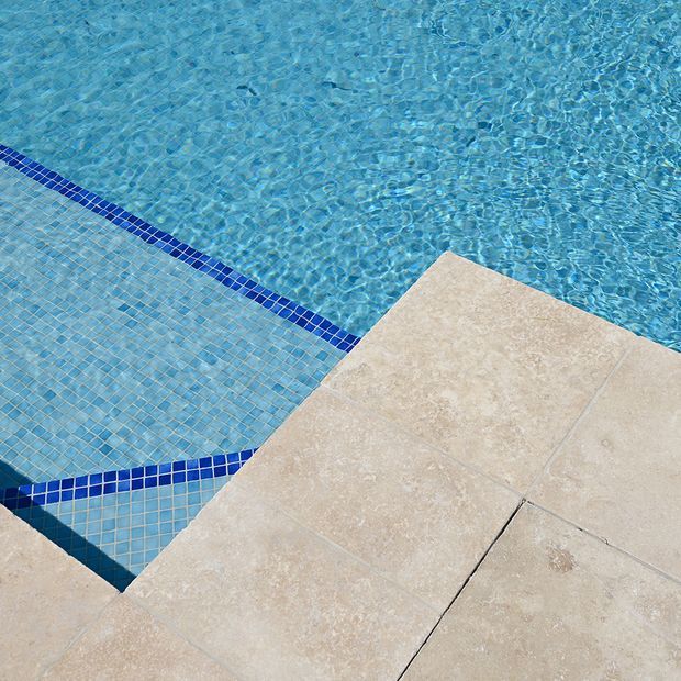 Close-up view of a swimming pool edge with blue mosaic-tiled surfaces, a dark blue tile border, clear turquoise water, and pale beige stone or porcelain paving around the pool.