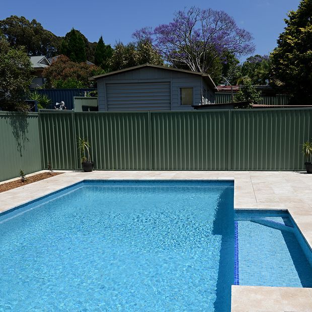 Sunny outdoor swimming pool area with a large rectangular blue pool and adjoining shallow section, surrounded by pale tiled paving and a tall green corrugated metal privacy fence. A small grey backyard structure and mature trees are visible beyond the fence.