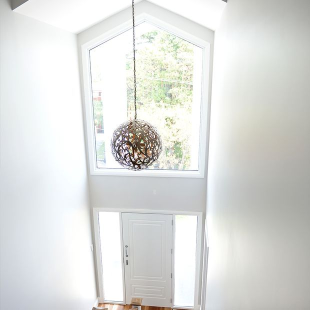 Bright white entryway viewed from above, featuring a tall vaulted space with a large polygonal window over a white front door and sidelights. A decorative spherical pendant light hangs centrally in front of the window.