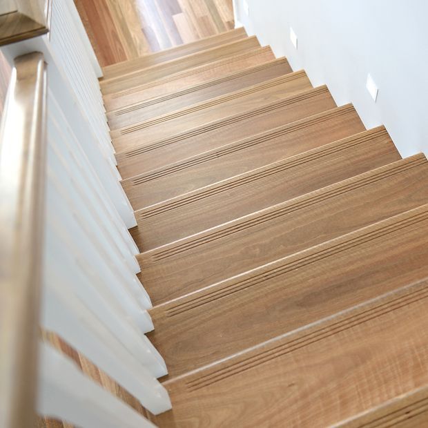 Close-up view looking up a straight interior staircase with warm wood-finished treads and matching timber flooring at the upper level. White painted walls and white vertical balusters create a bright, clean stairwell, with a timber handrail visible along the left side.