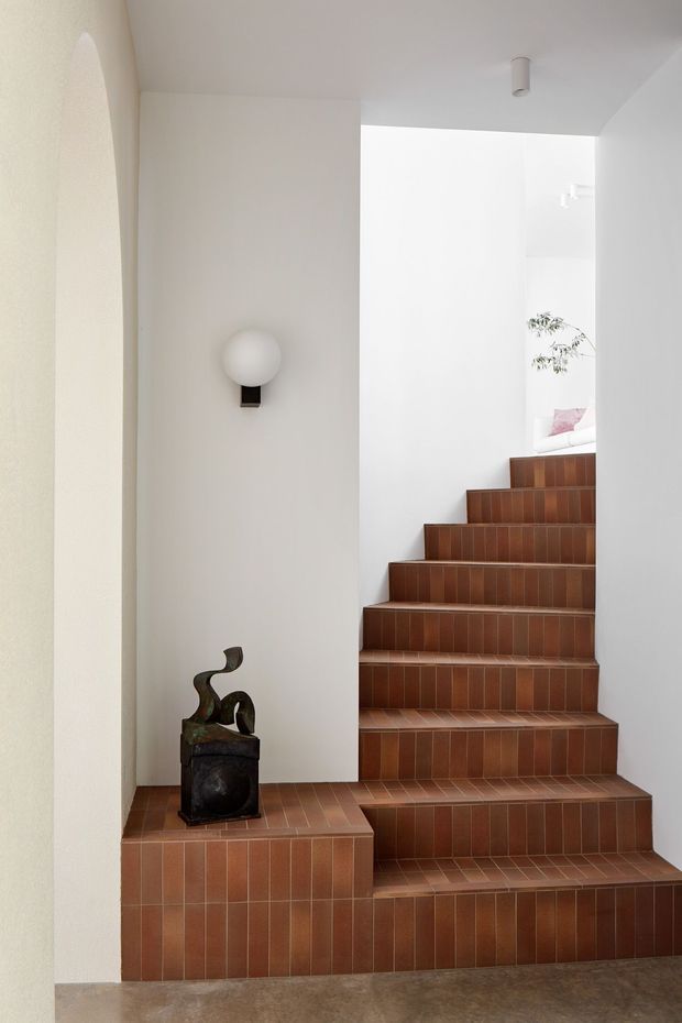 Bright minimalist stairway with warm terracotta-brown tiled steps and a tiled side plinth, enclosed by smooth white plaster walls. A simple globe wall sconce and small sculptural object provide restrained decorative detail, while an upper landing opens toward a partially visible bedroom area and plant.