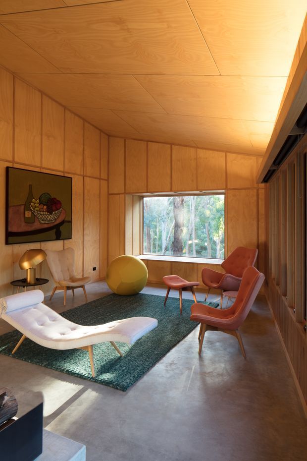 A warm, wood-panelled mid-century-inspired living room with a low sloping timber ceiling and full-height timber wall panelling. The room contains sculptural lounge chairs, a white chaise, a round yellow accent seat, a dark teal rug, a small side table, and a floor lamp. A large picture window looks onto trees, while vertical timber screening and glazing line the right side. The polished concrete floor and abundant natural light create a refined yet cosy atmosphere.