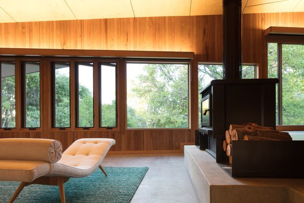 A warm, wood-lined living room with expansive dark-framed windows overlooking trees. The room features a sculptural upholstered sofa and chaise, a teal rug, a concrete floor, and a freestanding wood-burning fireplace with exposed logs on a substantial concrete hearth. The interior has a strong mid-century character, with vertical timber wall panelling, broad glazing, and a low horizontal composition.