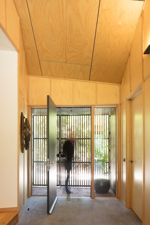 A narrow timber-lined entry hall with a high, angled plywood ceiling and matching panelled walls. The concrete floor leads to an open glazed door and a screened exterior entrance, while a timber door and built-in panel detailing line the right side. Warm natural wood dominates the space, with strong daylight filtering through vertical outdoor screening.