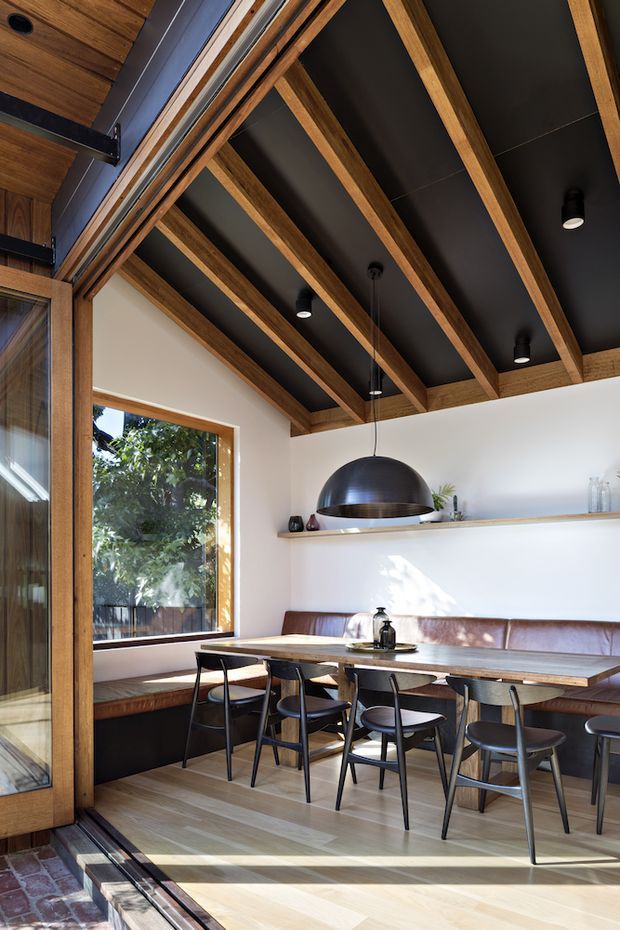 A warm, contemporary dining area with a long timber dining table, dark stools, and a built-in brown leather banquette beneath a floating timber shelf. Large windows and glazed doors bring in abundant daylight and views of greenery. The room is defined by exposed timber beams set against a dark ceiling, white walls, timber flooring, and a large black dome pendant light.