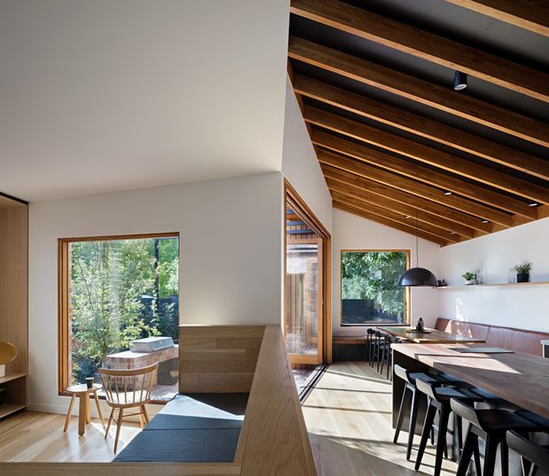 Open-plan contemporary interior with a dining and kitchen area beneath a dramatic exposed timber-beam ceiling. Warm timber flooring, cabinetry, window frames and dining furniture contrast with white walls and a long white kitchen island. Large windows overlook leafy greenery, while a raised timber-lined seating or living nook occupies the foreground.