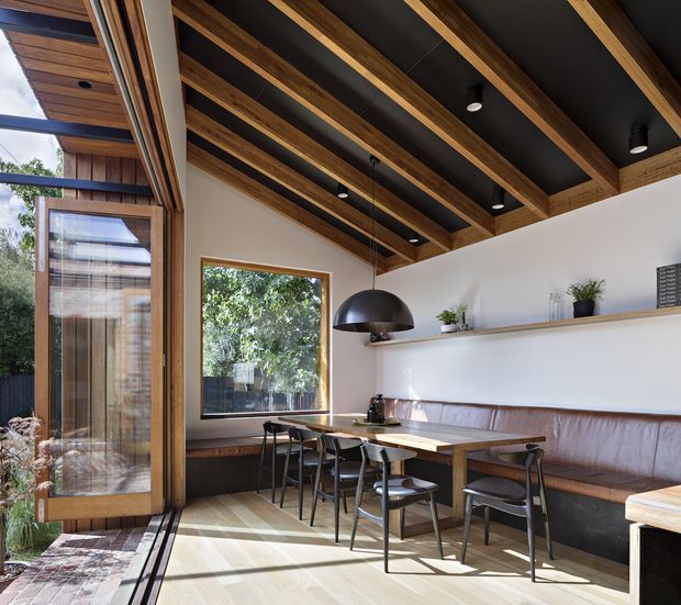 A warm, contemporary dining area with a long built-in leather banquette, dining table and chairs, and a large sculptural pendant light. Full-height timber-framed glazing opens to an exterior terrace, while exposed timber ceiling beams contrast with a dark ceiling lining. White walls, timber flooring, and a wall-mounted shelf with plants complete the refined interior.