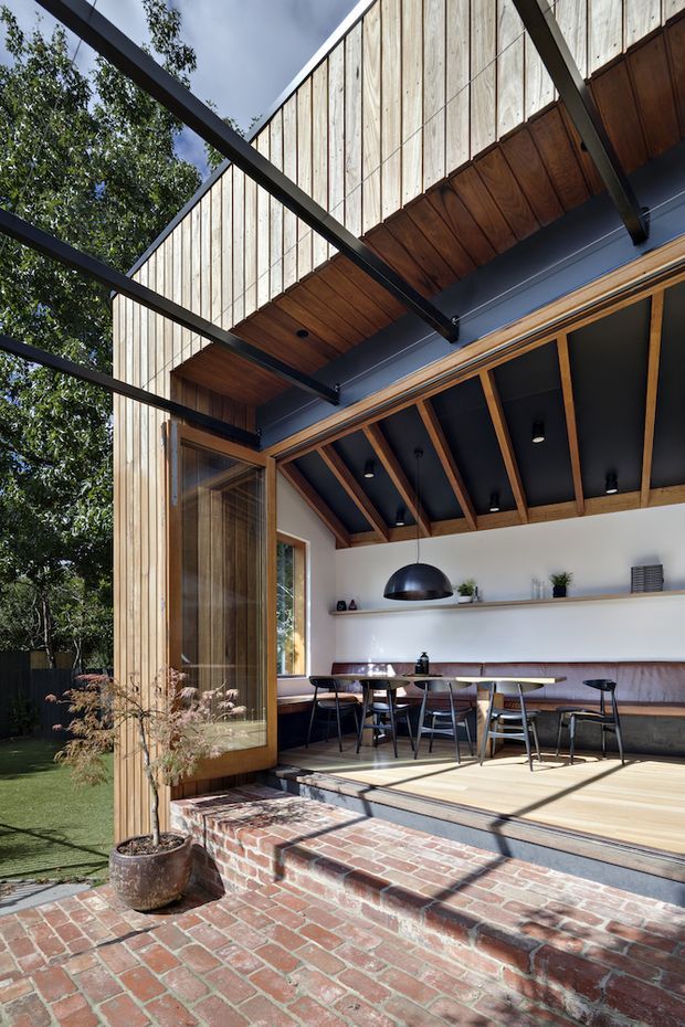 A bright indoor-outdoor dining and entertaining space viewed from a brick-paved courtyard. The open facade features extensive timber cladding and framing, large glazed doors, a covered deck, and a dramatic exposed-beam ceiling with dark painted panels and timber rafters. Inside are a long built-in banquette, dining tables and chairs, open shelving, pendant lighting, and potted plants. Black steel beams extend over the outdoor area, creating a strong contemporary architectural frame.
