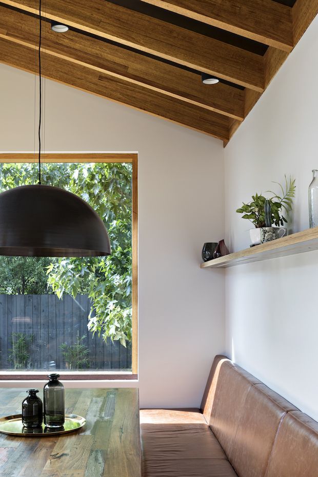 A bright, compact dining nook with a built-in brown leather banquette along a white wall, a rustic timber dining table, and a large dark dome pendant light. A broad timber-framed window overlooks leafy vegetation and a dark fence. Exposed timber ceiling beams with recessed downlights and a simple timber display shelf add warmth and texture.