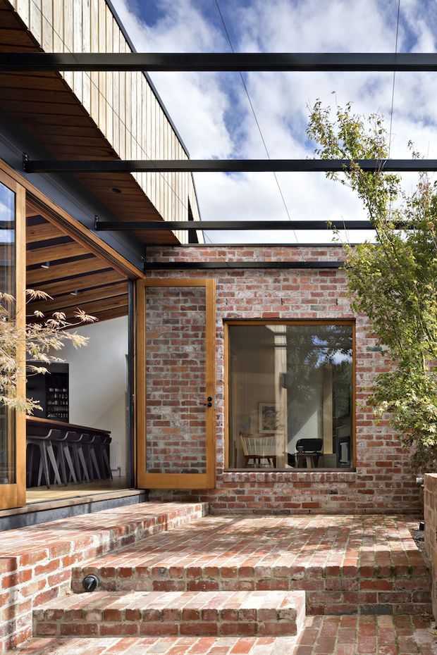 A sunlit brick courtyard beside a contemporary residential extension. The space features red and weathered brick paving and stepped seating, timber-framed glazing, a covered outdoor structure with exposed timber soffit and dark steel beams, and a partially visible interior bar area. Mature planting softens the courtyard edge.