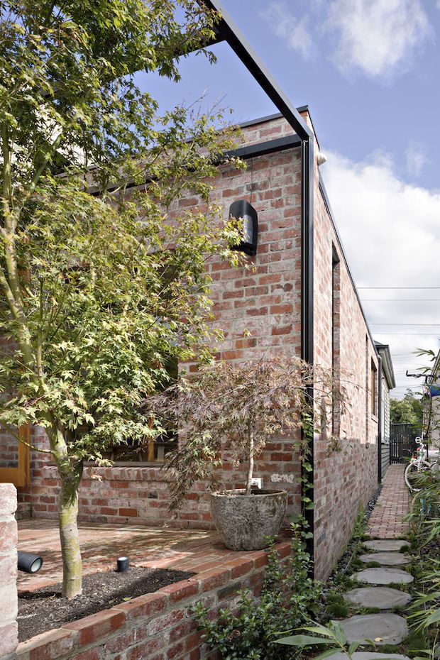Exterior view of a narrow brick-clad residential building with a simple dark metal roof edge and black-framed detailing. A landscaped brick courtyard sits beside the facade, with raised brick garden beds, mature planting, a large planter, and a stepping-stone path along the side of the building.