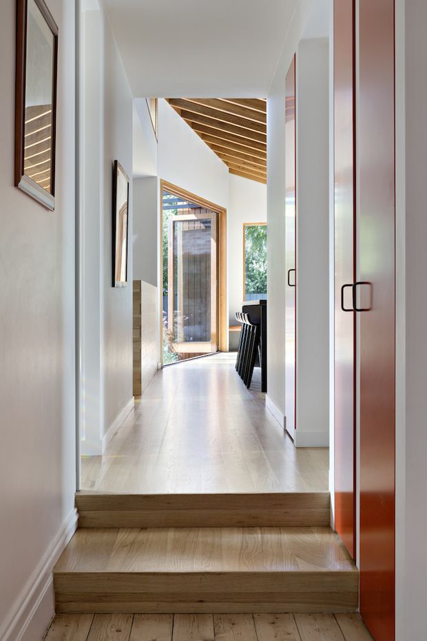 A bright, refined residential hallway viewed from a short flight of timber stairs. The corridor has pale timber flooring, white walls and ceiling, tall white columns, framed artwork, and glossy reddish-brown built-in cabinetry with dark handles. At the far end, a large glazed timber-framed door opens toward greenery, while exposed timber ceiling battens add warmth and architectural interest. A dark island or bench is partially visible beyond the corridor.