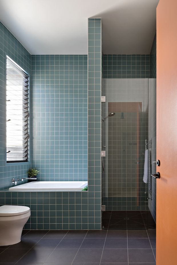 A compact contemporary bathroom with teal-blue square wall tiles, dark grey floor tiles, a white built-in bathtub, partial toilet, glass-enclosed shower, louvred window, and a warm orange door. A tiled partition separates the bathtub from the shower area.