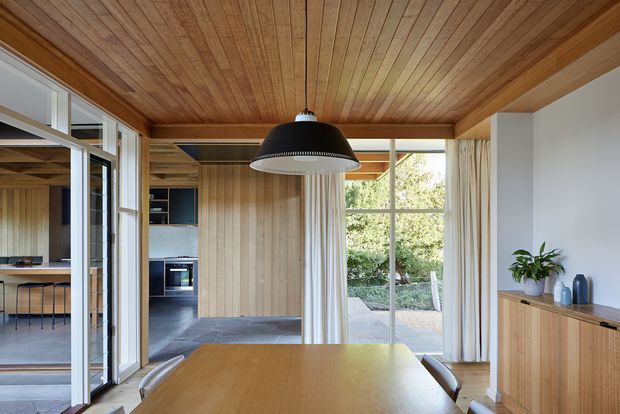 A warm, timber-lined dining room with a large rectangular wood dining table and pendant light, opening directly toward a compact kitchen and a glazed garden-facing outdoor area. Full-height timber ceiling and vertical timber wall panelling create a cohesive natural interior, with built-in timber cabinetry along the right wall and extensive framed glazing and sliding doors.