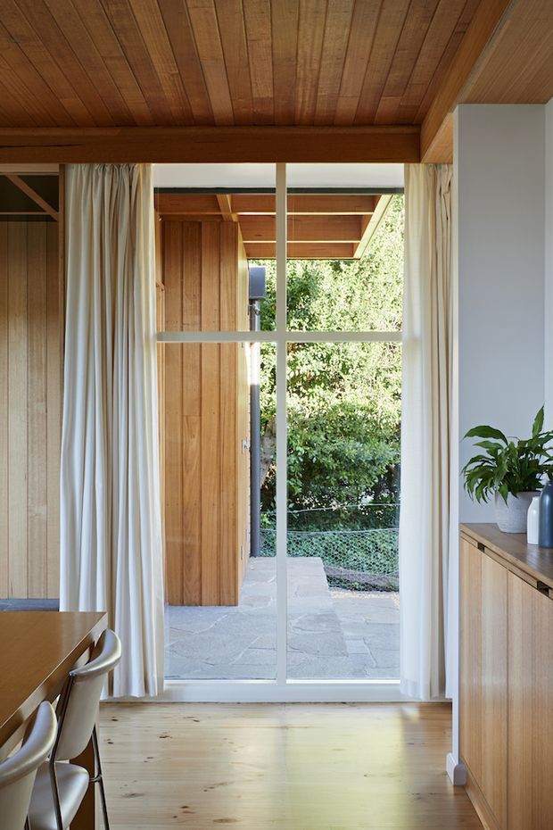 A warm, timber-lined interior viewed toward a full-height glazed door or window opening onto a paved outdoor area and leafy garden. The image shows a dining area at left with a timber table and chairs, light curtains framing the glazing, pale timber flooring, and a fixed timber sideboard or cabinet with a potted plant at right. The ceiling and wall panelling are predominantly natural timber, creating a cohesive, calm contemporary interior with strong indoor-outdoor connection.