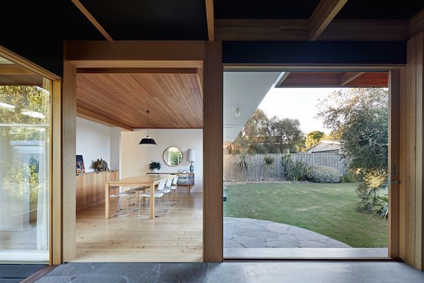 View from a glazed interior threshold into a light-filled dining area and landscaped backyard. The room features extensive timber framing, a warm timber-lined ceiling, pale timber flooring, white walls, a timber dining table with white chairs, and large sliding glass openings connecting directly to a lawn and stone-paved outdoor area. Dark ceiling framing provides contrast, while the garden is visible beyond the glazing.