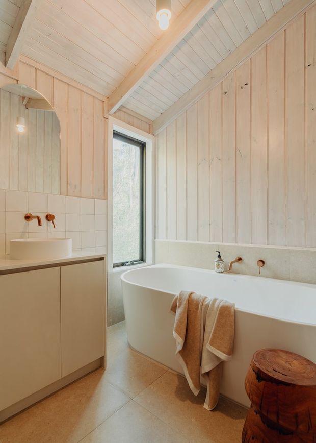 A warm, light-filled bathroom with pale timber wall panelling and a vaulted, timber-lined ceiling. The room features a freestanding white bathtub, a tiled vanity with vessel basin, brushed copper or bronze wall-mounted tapware, a tall window, and a natural timber stool. The design is simple, organic, and spa-like.