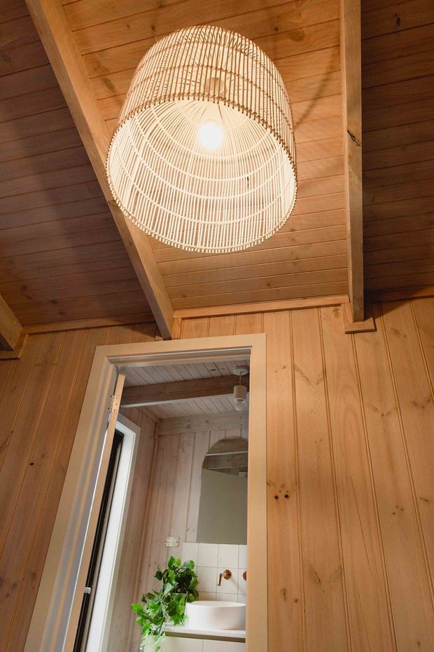 Warm timber-lined interior with exposed timber ceiling beams and vertical wall panelling. A large woven pendant light hangs centrally above a doorway leading into a compact bathroom, where a white basin, tiled splashback, mirror, and indoor plant are visible.