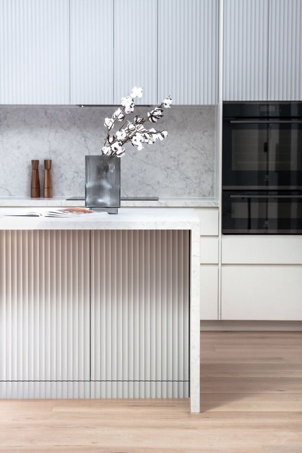 A bright, refined contemporary kitchen centred on a large island with a vertically fluted white-panelled base and pale stone benchtop. The rear wall features a veined stone splashback, integrated cabinetry, tall black built-in ovens, and vertically ribbed light-coloured wall panelling. Pale timber flooring and restrained styling create a clean, softly luxurious look.