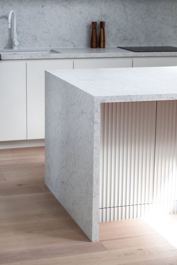 Close-up architectural interior photograph of a minimalist kitchen island clad in pale veined stone, with a fluted vertical-panel detail on one side. The background shows white flat-panel cabinetry, a matching stone benchtop and splashback, an undermount sink with a slim light-coloured tap, and warm timber flooring. A pair of dark timber vessels provide a small accent on the rear benchtop.