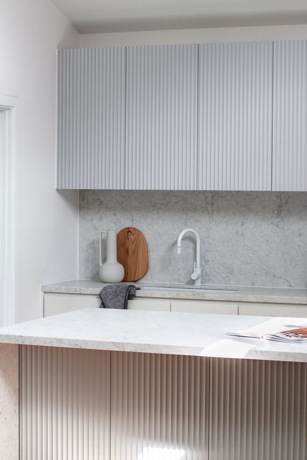 Bright, refined contemporary kitchen featuring a large marble-look island with vertically fluted cabinetry, matching fluted overhead cupboards, a pale stone splashback and benchtop, and a minimalist white gooseneck tap. Warm timber accessories and a dark textured tea towel provide subtle contrast.
