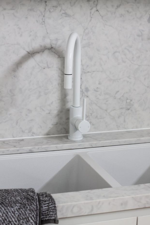 Close-up of a white gooseneck kitchen faucet mounted above a large white double-bowl sink. The splashback, benchtop, and surrounding surfaces are clad in pale grey-white veined marble, with white cabinetry below and a dark grey textured towel draped over the sink edge.