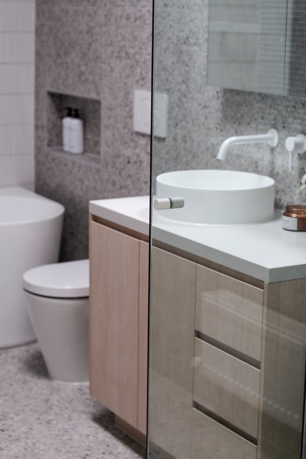 A compact contemporary bathroom viewed through a clear glass shower screen, featuring a freestanding white toilet, a round vessel basin on a white benchtop, light timber vanity cabinetry, grey terrazzo-like wall and floor surfaces, and a recessed wall niche with toiletries. A large mirror reflects the vanity and wall-mounted tapware.