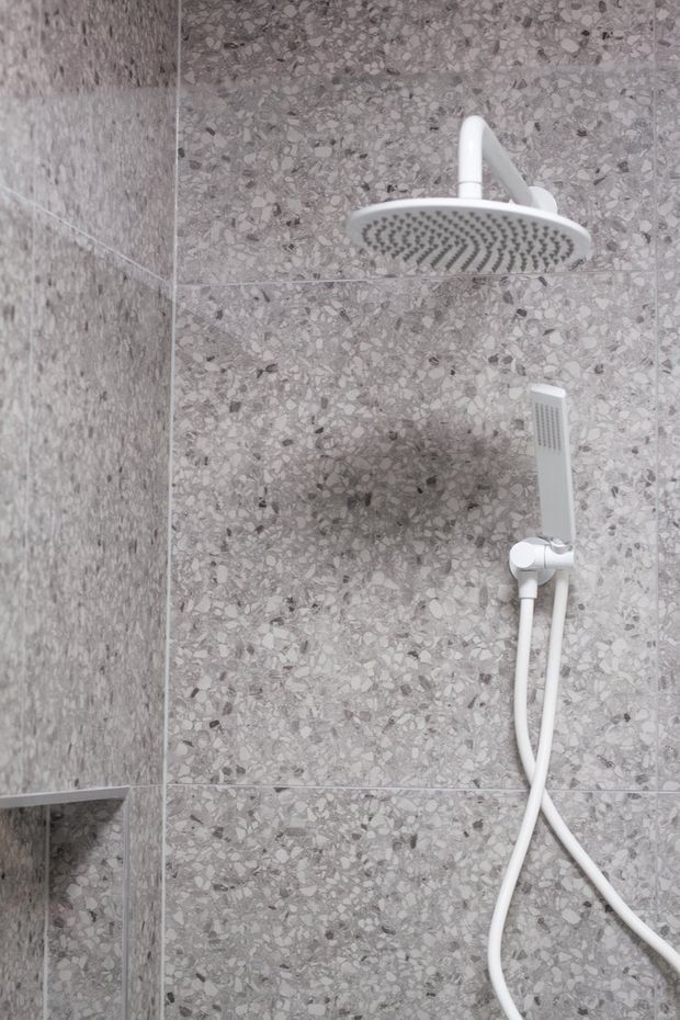 Close-up view of a modern shower installation with a white round rain showerhead and matching handheld shower on a grey terrazzo-look tiled wall. The shower area has clean contemporary detailing and a neutral grey palette.