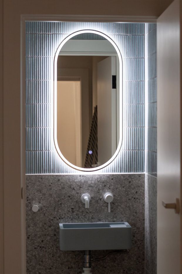 Compact contemporary bathroom washbasin area featuring a wall-mounted grey basin, exposed chrome tapware, a large rounded-rectangle illuminated mirror, vertically ridged light blue-grey wall tiles, and a dark speckled stone or terrazzo splashback. A doorway and hallway are visible in the mirror reflection.