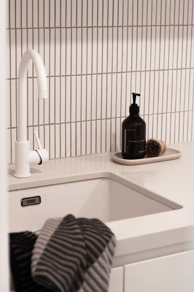 Close-up of a clean white kitchen sink area with a white gooseneck tap, integrated basin, pale benchtop, white cabinetry, and a vertical white tiled splashback. A dark soap dispenser and brush sit on a small tray beside the sink, with a striped cloth draped over the cabinet front.