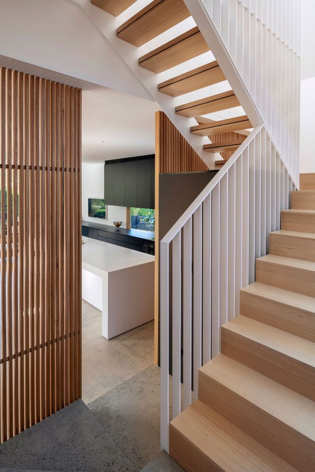 A bright contemporary stairway viewed from an open-plan kitchen area. The staircase has light timber treads with open risers, white stringers, and slim white vertical balustrades. Vertical timber battens screen the kitchen and create a warm architectural feature, while dark matte cabinetry and a white kitchen island are visible beyond. The floor appears to be polished concrete or terrazzo-like concrete, and the space has a clean, refined, natural-material palette.