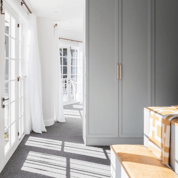 Bright, narrow interior passage or dressing area with tall white French doors and sheer curtains on the left, a large built-in grey wardrobe with brass handles on the right, and a small upholstered bench in the foreground. Strong sunlight casts geometric window shadows across the grey carpet, creating an airy, refined atmosphere.