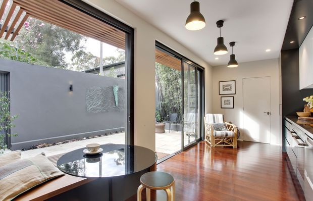 Bright contemporary open-plan interior with a compact kitchen, round dining table, timber flooring, extensive sliding glass doors, and a leafy courtyard beyond. A timber slatted overhead screen shades the outdoor edge, while black pendant lights and dark kitchen cabinetry provide contrast against pale walls.