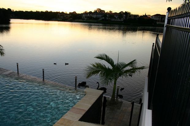 A waterfront outdoor area viewed from an elevated position, featuring a swimming pool in the foreground beside a calm lagoon or lake. A palm tree, pool safety fencing, tiled paving, and a dark vertical-slat building facade frame the scene, with residential buildings visible across the water at sunset.