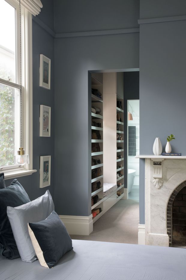 A refined blue-grey bedroom with a large sash window, upholstered bed, layered blue and white cushions, framed artwork, ornate white fireplace, and a doorway leading to a built-in walk-in wardrobe. The room has traditional detailing, including decorative cornices, skirting boards, and a carved fireplace surround, with soft natural light and a calm, muted palette.