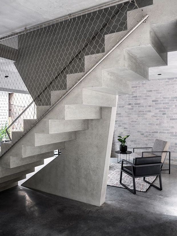 Interior view of a sculptural concrete stairway with cantilevered treads and a chain-link safety balustrade. The stair sits within an industrial-style living area with polished concrete flooring, a pale exposed brick wall, and a black-framed lounge chair beside a small table and plant.