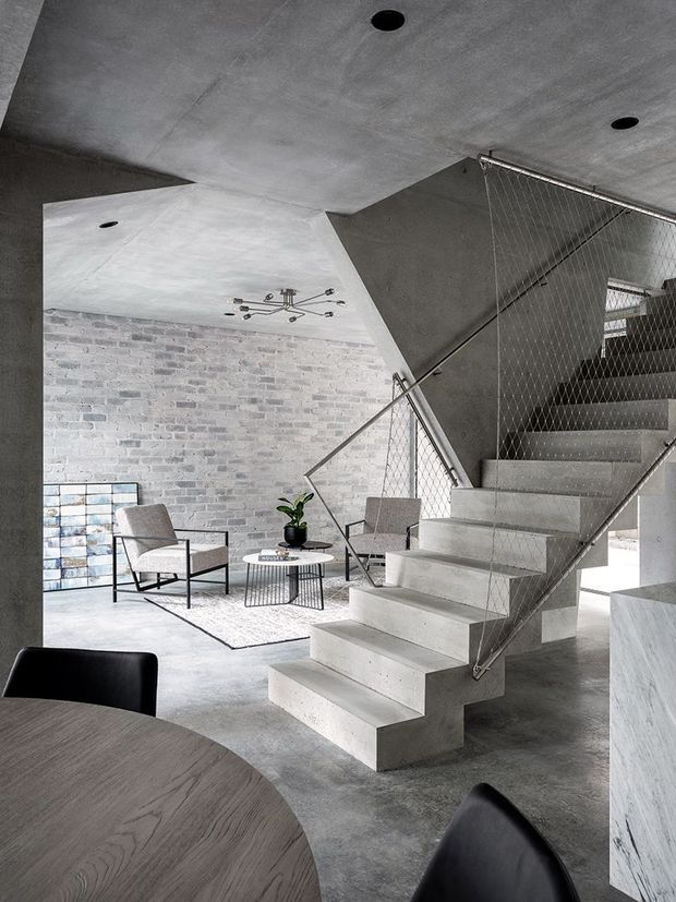 A contemporary industrial interior featuring a sculptural concrete staircase with a wire-mesh balustrade, exposed grey concrete ceilings and floors, and a pale exposed-brick feature wall. A small seating area with neutral armchairs, a round coffee table, and a geometric wall artwork sits beneath the stair. The foreground includes a dark timber dining table and black chairs, while minimalist black track-style ceiling lights and a potted plant add restrained detail.