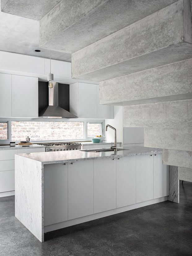 A contemporary kitchen with a large white island, integrated cabinetry, stone benchtops, stainless steel tapware and appliances, and a long horizontal window overlooking exposed brickwork. The space features dramatic exposed concrete ceiling beams and a polished industrial material palette of pale cabinetry, grey concrete, and dark flooring.