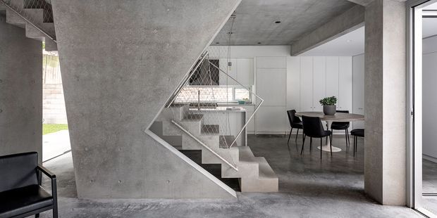 Contemporary open-plan interior featuring a sculptural concrete staircase with metal mesh balustrades, polished concrete floors, exposed concrete structure, and a minimalist dining area with white built-in cabinetry. Large glazed openings bring in natural light and connect the interior to the outdoors.