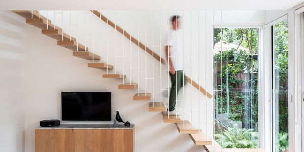 Bright contemporary interior featuring an open-riser timber staircase with slender white vertical cable balustrades, white panelled walls, and a timber media console beneath a wall-mounted television. Full-height glazing at right opens to a leafy garden, while a person is visible ascending the stairs.