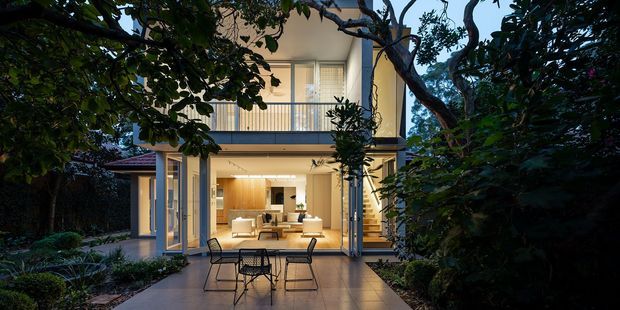 A contemporary two-storey home viewed from a landscaped outdoor terrace at dusk. Large glazed openings reveal a warmly lit open-plan kitchen and living area, while an upper-level balcony with slim metal balustrading sits above. Dense mature planting frames the facade and terrace.