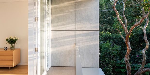A refined interior transition beside a garden, featuring a pale timber sideboard with a vase and clock, a tall glazed opening, and a minimalist grey stone or concrete-clad wall with integrated cabinetry and a projecting bench. Lush trees and planting are visible outside.