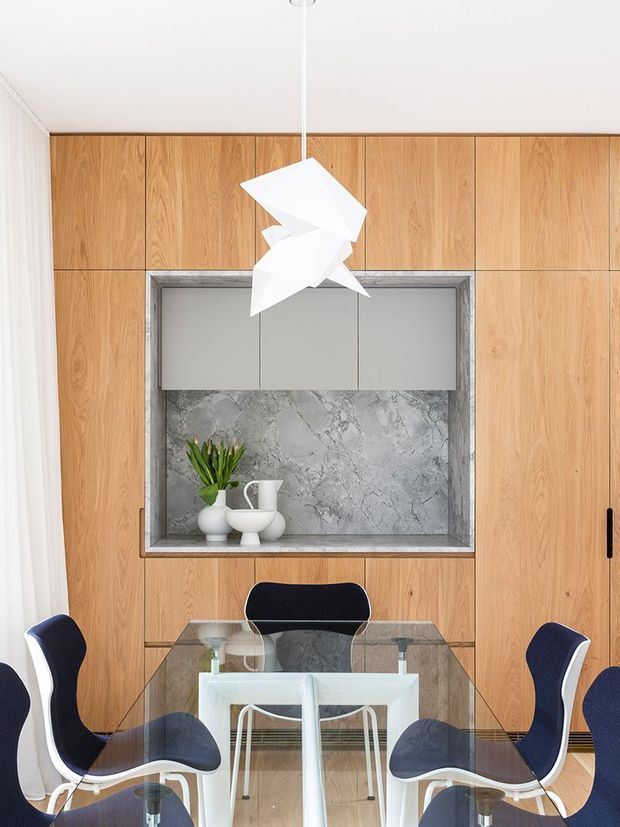 Refined dining area with a glass dining table and sculptural white dining chairs, set against a full-height warm timber-panelled wall. A recessed display niche features a grey marble-look back panel, minimalist grey cabinetry, white ceramics and a small plant. A geometric white pendant light hangs centrally above the table, creating a clean, contemporary composition.