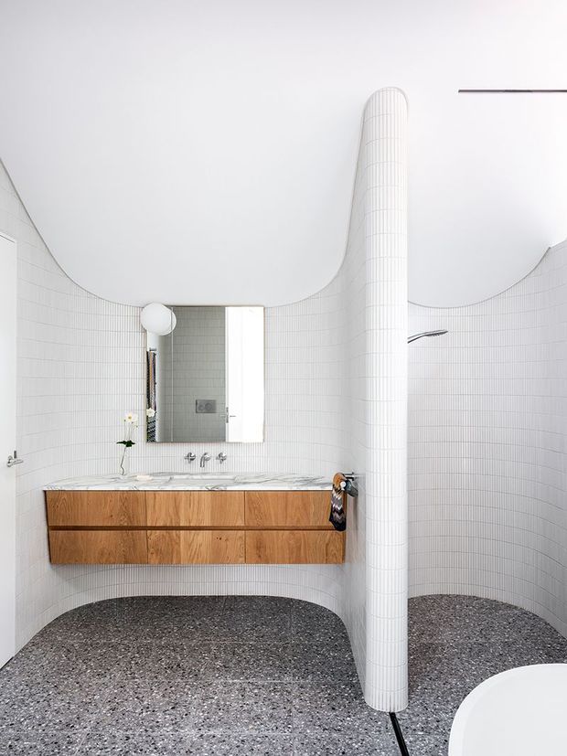 A sculptural contemporary bathroom with curved white tiled walls and a rounded tiled partition column. A floating timber vanity with a white marble benchtop sits beneath a mirror, while the floor is finished in dark terrazzo. The space has a bright, minimal, refined appearance with soft curved forms and partially visible bath and shower fittings.