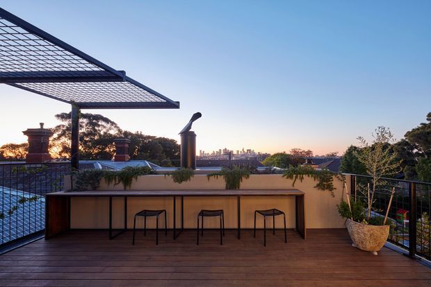 A rooftop terrace at dusk with timber decking, a built-in outdoor bar or counter with stools, planter boxes and potted greenery, black metal balustrades, and a large open steel pergola overhead. The terrace overlooks surrounding rooftops and a distant urban skyline.